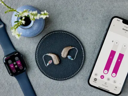 Still-life com Apple Watch, aparelhos Oticon Intent e iPhone mostrando o app Oticon Companion com controles de volume L/R